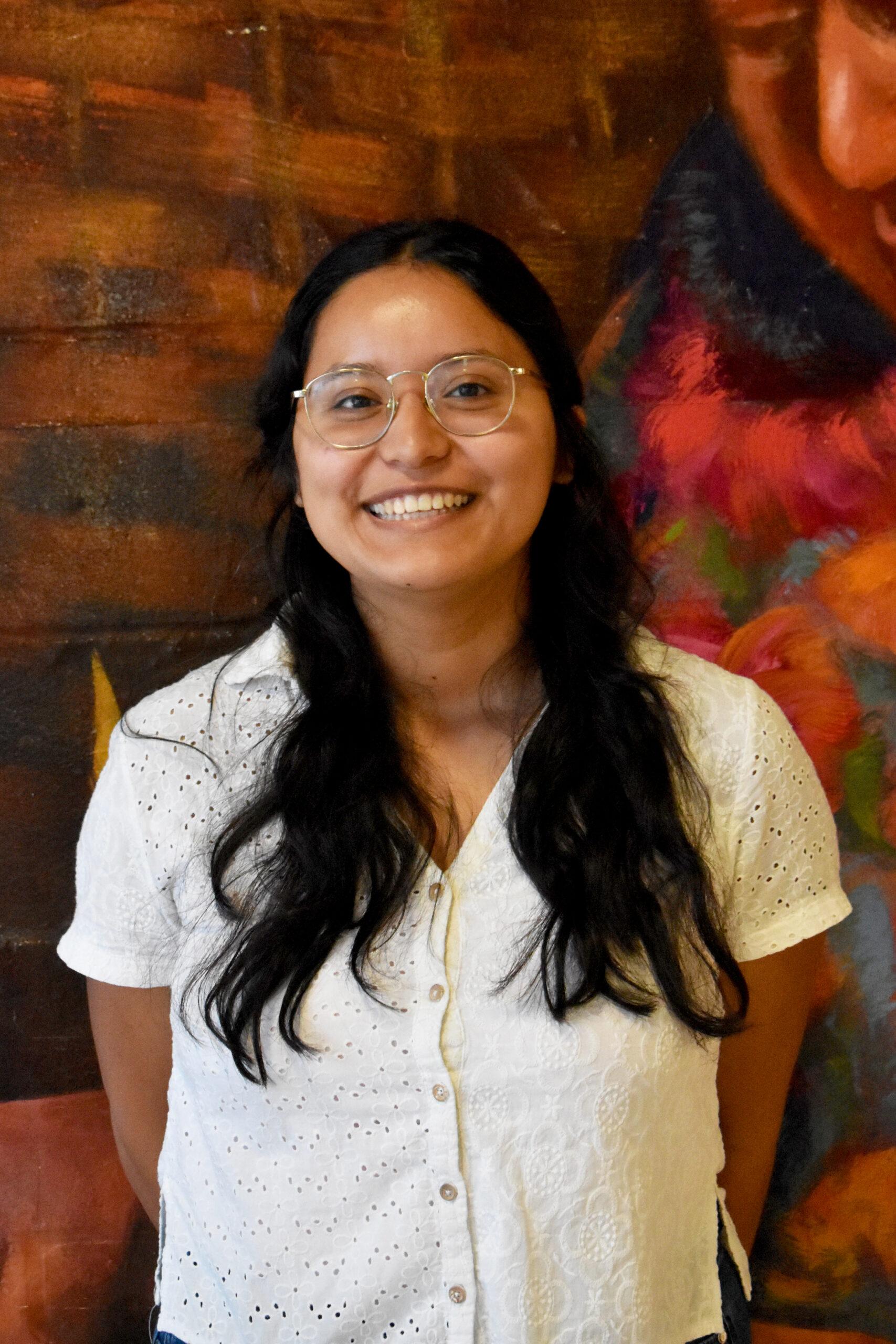 A smiling young woman wearing a white button-down shirt looks straight ahead at the camera.