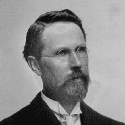 Silas Mason poses for the camera wearing round, thin-framed glasses, a white dress shirt and a black coat.