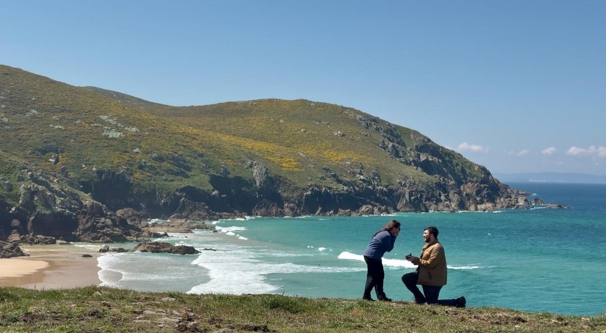 A kneeling man proposes to a woman in a grassy area near the beach, with bright blue waves rushing toward the sandy shore in the background.