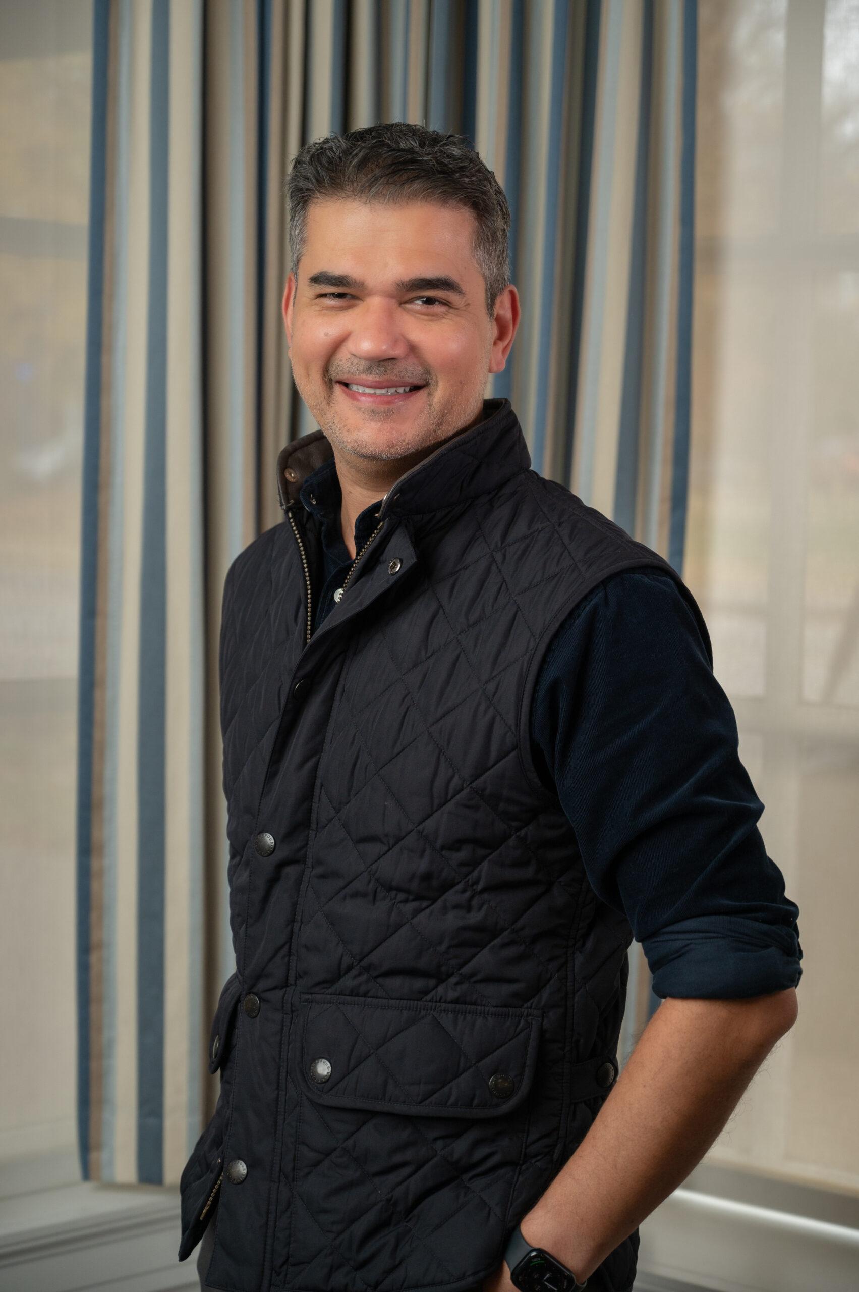 Ajay Nanda stands in front of a gray background. He is smiling at the camera with his hands in his pockets and is wearing a black shirt and puffy vest.