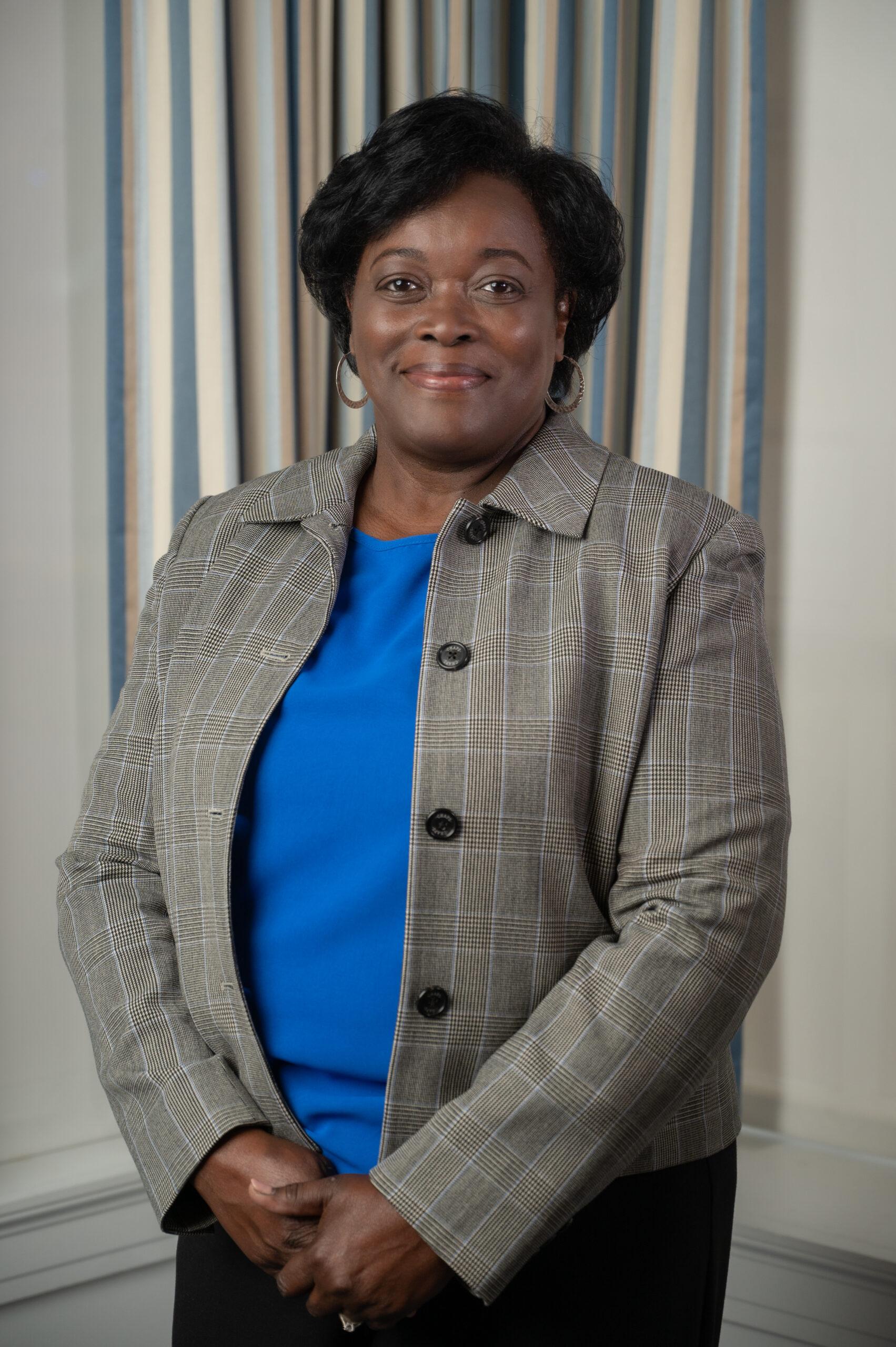 Brenda Lane stands in front of a gray background. She is wearing a gray jacket and a Berea blue shirt.
