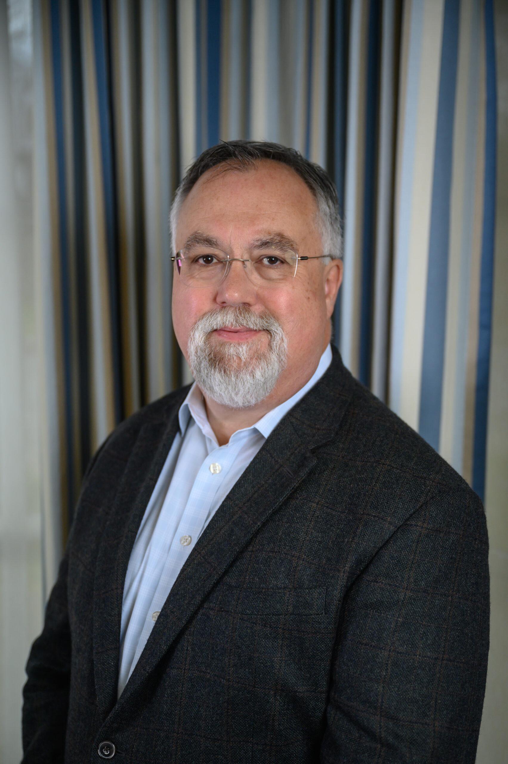 John Graham stands in front of a gray backdrop. He wear glasses and has on a white shirt and black blazer.