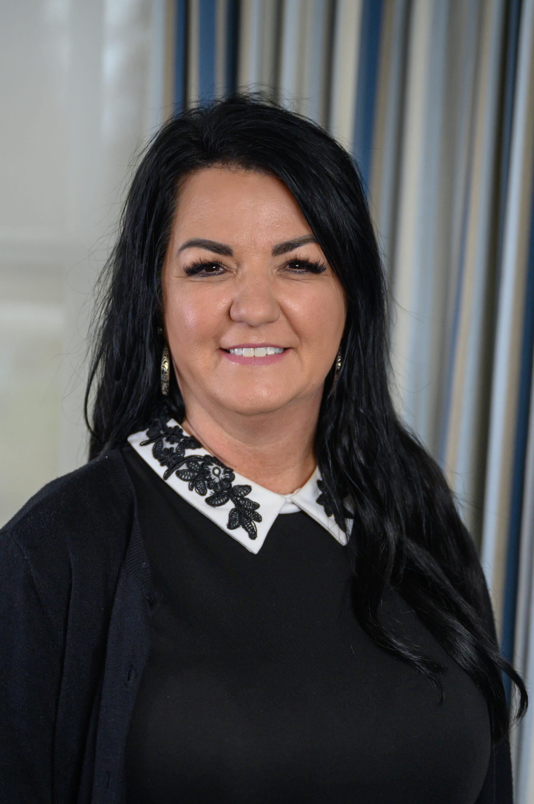 Vestena Robbins stands in front of a gray background. She is wearing a black top with a white beaded collar. She has black hair and is smiling at the camera.